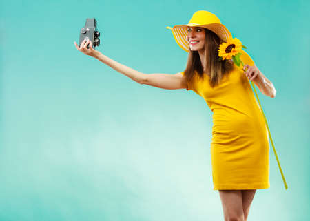 summer woman wearing yellow dress and hat with sunflower taking self picture with old vintage camera on vivid blue backgroundの写真素材