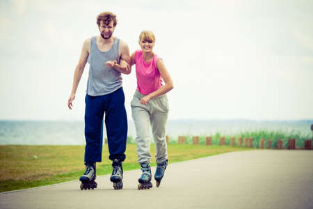 Active holidays, exercises, relationship concept. Young woman and man dressed up in sporty way, holding their hands while rollerblading together on promenade.の写真素材