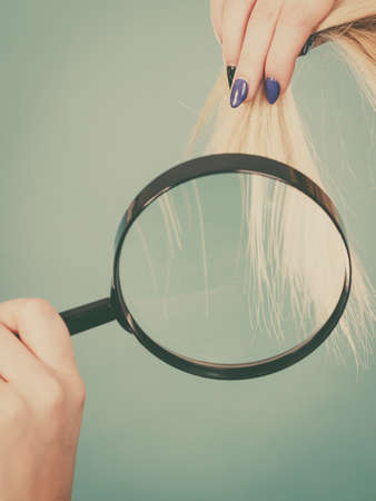 Haircare and hairstyling, bad effects of bleaching concept. Blonde woman looking at her damaged, split hair ends through magnifying glass.の写真素材