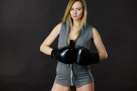 Young hooded woman fighting boxing. Blonde attractive girl wearing black punch gloves. Sport and...
