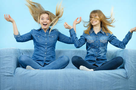 Two funny shocked women wearing jeans shirts having windblown blonde hair. Blue background.の写真素材
