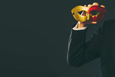 Holidays, people and celebration concept. Male hand holding red golden carnival mask closeup on darkの写真素材