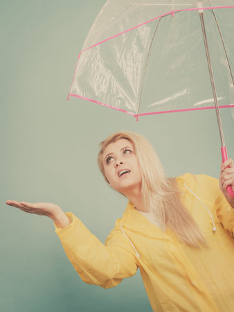 Blonde woman wearing yellow raincoat holding transparent umbrella checking weather if it is raining.の写真素材