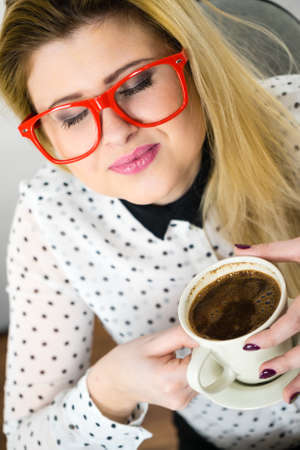 Happy young woman wearing eyeglasses at office drinking hot coffee or tea enjoying her break time during work. Job relax.の写真素材