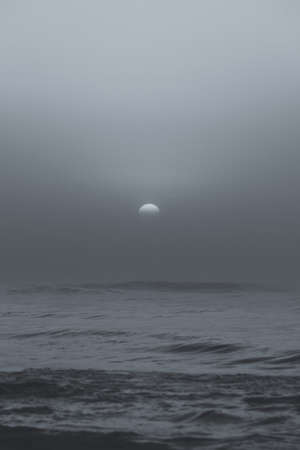 Black and white shot of sun behind fog or clouds during sunset by beach.の写真素材