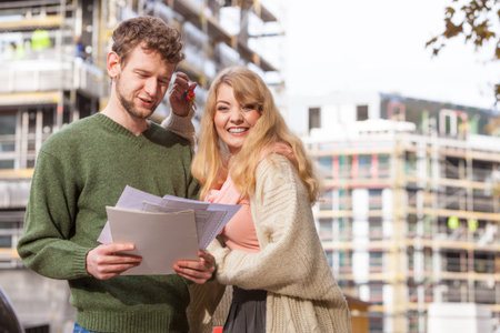 Real estate and family concept - young couple on front of new big modern house construction site with blueprint project keys dreaming about homeの写真素材