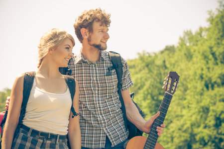 Adventure, tourism, enjoying summer time together - Hiking young couple with guitar backpack tramping on forest road sunny countrysideの写真素材