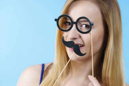 Woman holding carnival accessories, eyeglasses and moustache on stick having fun. Studio shot on blue background.の写真素材