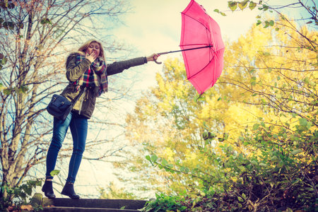 Autumn weather concept. Woman walking in park holding red umbrella fighting with strong wind