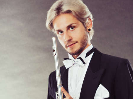 Classical music, passion and hobby concept. Portrait of elegantly dressed musician blonde young man holding flute. Studio shot on dark grey backgroundの写真素材