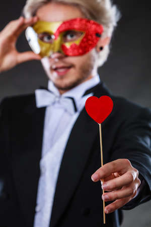 Valentines day. Love concept. Holidays and celebration. Elegant young guy wearing suit white shirt bow tie with carnival mask and heart stick in hand, on dark.の写真素材