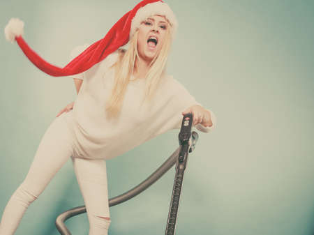 Woman vacuuming the house, last minute. Teen girl in santa helper hat with vacuum cleaner, funny face expression. Christmas time and housework concept.の写真素材