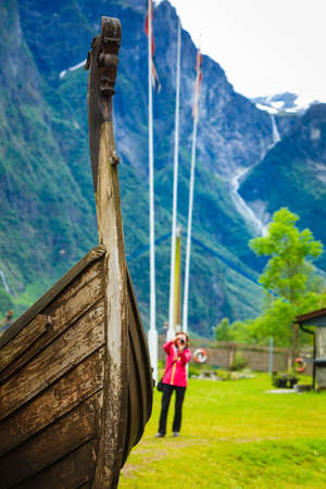 Female tourist standing near old wooden viking boat in norwegian nature, taking photo with camera. Tourism and traveling conceptの写真素材