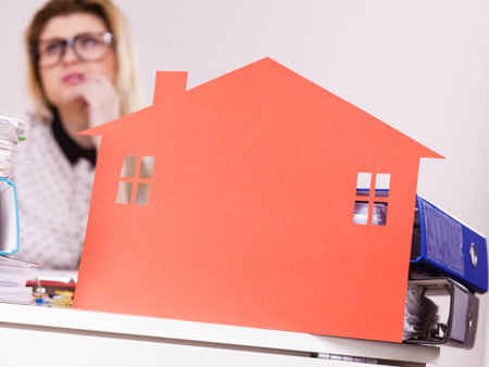 Thinking business woman sitting working at desk full off documents in binders, she having big red house made of paper, real estate concept.の写真素材
