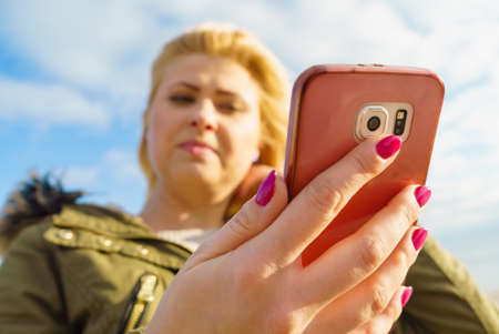Social media, technology, modern devices, internet concept. Woman teenager wearing warm coat using her smartphone while walking outside on sunny day, sky with clouds in backgroundの写真素材