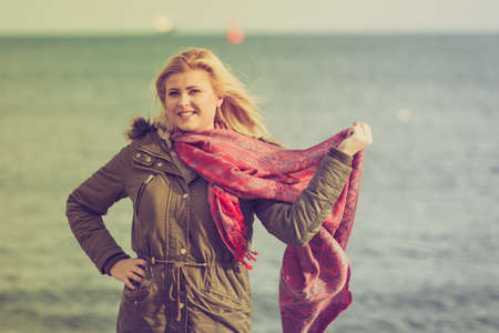 Happiness, enjoying weather, fashion concept. Woman wearing warm coat holding red beautiful scarf spending her time outside on the beachの写真素材