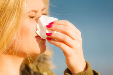Seasonal flu, cold, runny nose concept. Sneezing blonde young woman into tissue, outside sunny winter shotの写真素材