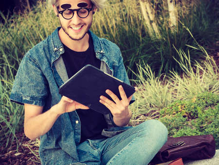Male fashion, technology, student concept. Guy with tablet wearing jeans outfit and eccentric sunglasses sitting on white ledgeの写真素材