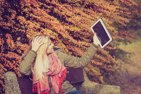 Woman relaxing sitting on bench in park during autumn weather using tablet pc taking selfie photo