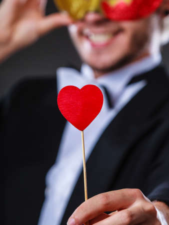 Valentines day. Love concept. Holidays and celebration. Elegant young guy wearing suit white shirt bow tie with carnival mask and heart stick in hand, on dark.の写真素材