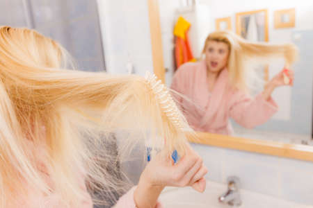 Woman wearing dressing gown trying to brush her long blonde very tangled hair, morning beauty routine. Haircare problem and hairstyling concept.の写真素材