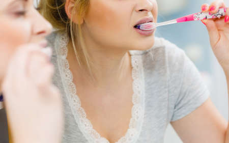 Woman brushing cleaning teeth closeup. Blonde girl with toothbrush in bathroom, looking at mirror. Oral hygiene.の写真素材