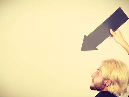 Planning, directions, choices concept. Man holding black arrow pointing left down. Indoor shot on light backgroundの写真素材