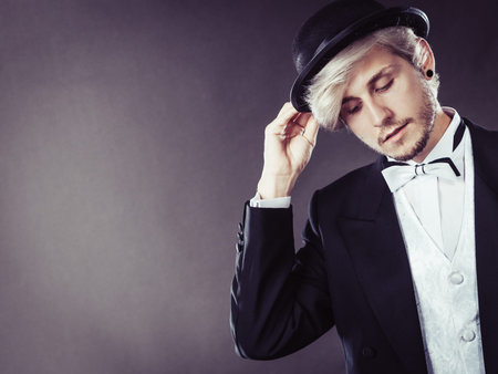 Tuxedo, male fashion, classical look concept. Elegantly dressed blonde man holding black fedora hat....
