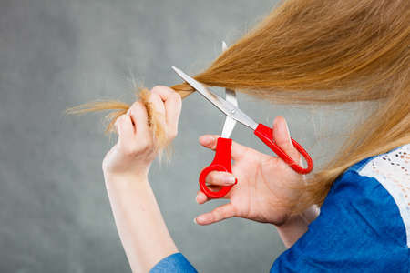 Cutting coiffure and new look. Part body blonde woman cut her long straight hair. Female hands with red scissors making modern hairstyle.の写真素材