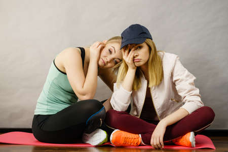 Two attractive women wearing sports clothes sitting on exercise mat being bored or tired after hard workout.の写真素材