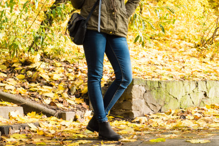 Woman legs standing in autumnal park during autumn weather, gold seasonal leaves on ground.