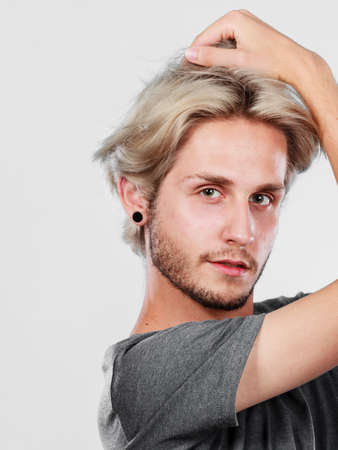 Hairstyle, haircare concept. Blonde handsome guy wearing grey t shirt touching his hair, studio shot grey background.の写真素材