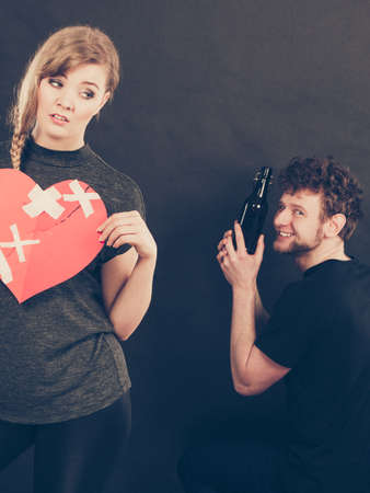 Unhappy couple. Family and alcoholism problems. Addiction and trouble of drinking. Man with alcohol bottle. Sad woman holds paper broken heart.の写真素材