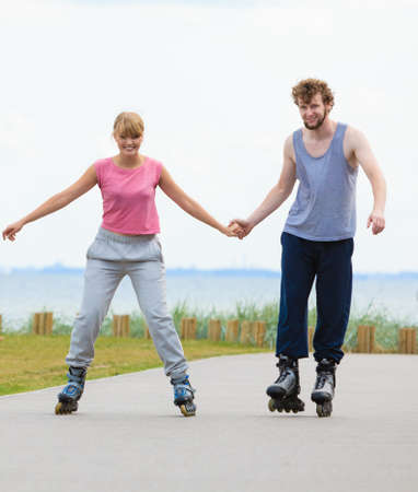 Active holidays, exercises, relationship concept. Young woman and man dressed up in sporty way, holding their hands while rollerblading together on promenade.の写真素材