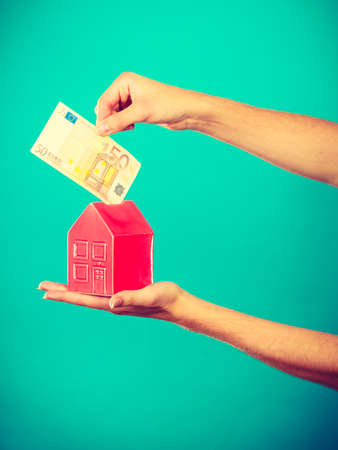 Household savings and finances, economy concept. Preson puts money into a piggy bank in the shape of a house, studio shot on blue backgroundの写真素材