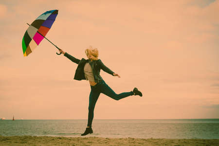 Happiness, enjoying cold autumn weather, feeling great concept. Woman jumping with colorful umbrella on beach near sea, sunny day and clear blue skyの写真素材