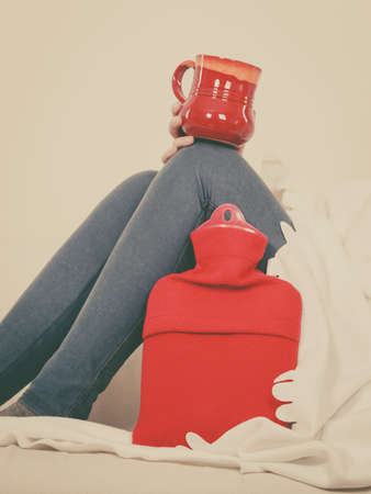 Woman having flu showing ideas how to deal with cold, holding hot water bottle and tea in cup sitting on sofaの写真素材