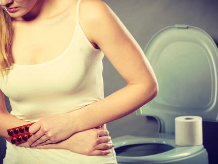 Bellyache, constipation or food poisoning. Woman suffering from strong abdominal pain, holding medical tablets activated carbon in hand, paper on toilet in the backgroundの写真素材