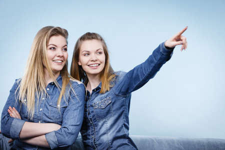 Friendship, human relations concept. Two happy women friends or sisters wearing jeans shirts sitting on sofa pointing somewhere.の写真素材