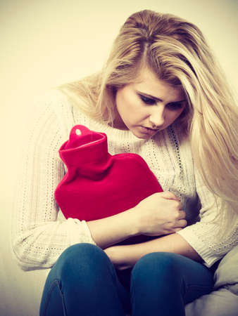 Painful periods and menstrual cramp problems concept. Woman having stomach cramps sitting on cofa feeling very unwell holding hot water bottle to feel some reliefの写真素材
