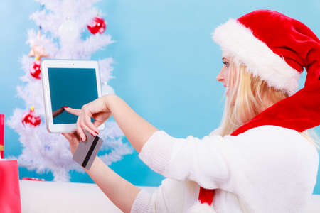 Online shopping, buying Christmas gifts on Internet concept. Woman in Santa hat sitting on sofa holding tablet and credit card, browsing net.の写真素材