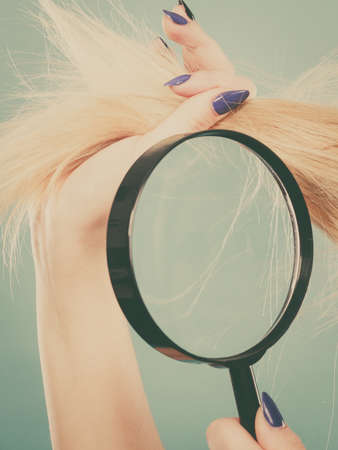 Haircare and hairstyling, bad effects of bleaching concept. Blonde woman looking at her damaged, split hair ends through magnifying glass.の写真素材