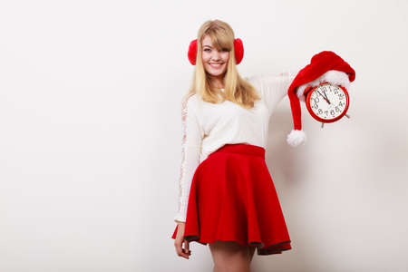 Happy cute woman holding alarm clock with santa claus hat. Pretty girl in earmuffs on gray. Christmas time season concept.の写真素材