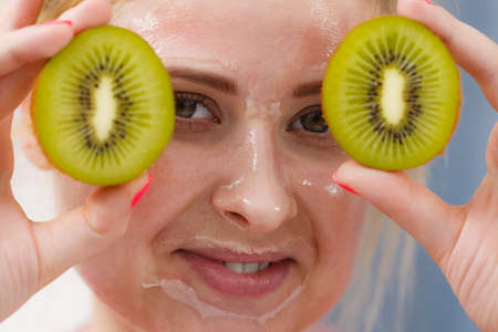 Facial skin and body care, vitamins good complexion treatment at home concept. Young woman having gel peel off mask on her face holding kiwi fruit.の写真素材