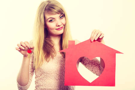 People, fresh start, real estate and love concept. Young enjoyable enamoured woman holding red house with big heart and keys to her future.の写真素材