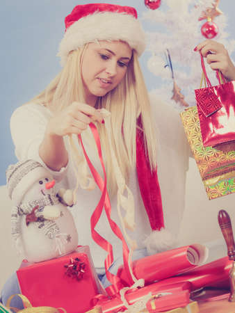 Christmas time concept. Happy teen blonde girl wearing santa claus hat preparing gifts for xmas, sitting at home on couch, cozy holiday interiorの写真素材
