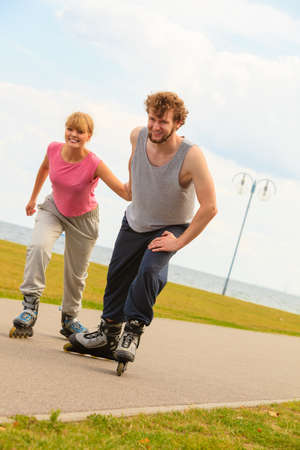 Active holidays, exercises, relationship concept. Young man dressed in sports clothes putting his girlfriend up to do rollerblading while holding her hand on promenadeの写真素材