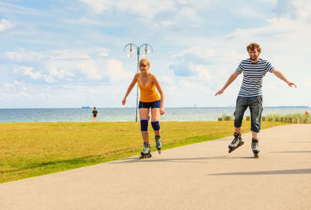 Holidays, active people and friendship concept. Young fit couple on roller skates riding outdoors on sea shore, woman and man rollerblading together on the promenadeの写真素材