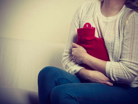 Painful periods and menstrual cramp problems concept. Woman having stomach cramps sitting on cofa feeling very unwell holding hot water bottle to feel some reliefの写真素材