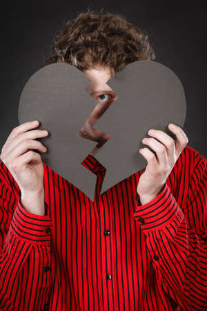 Love romance relationship heartbreak unhappy sadness shame concept. Man covering behind cutout. Young male hiding his face with parts of broken heart.の写真素材